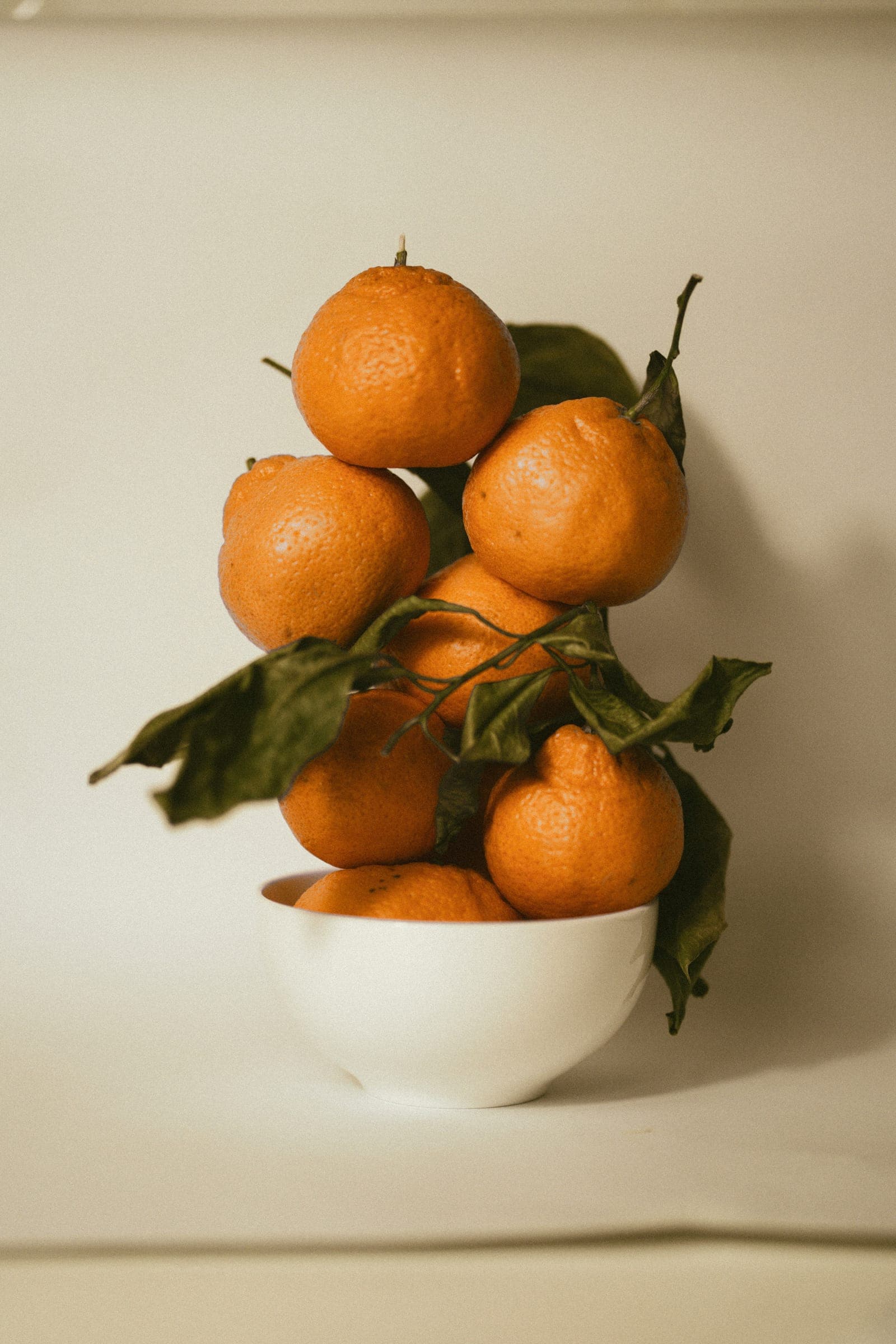 A bowl of fresh oranges with leaves on a warm neutral background